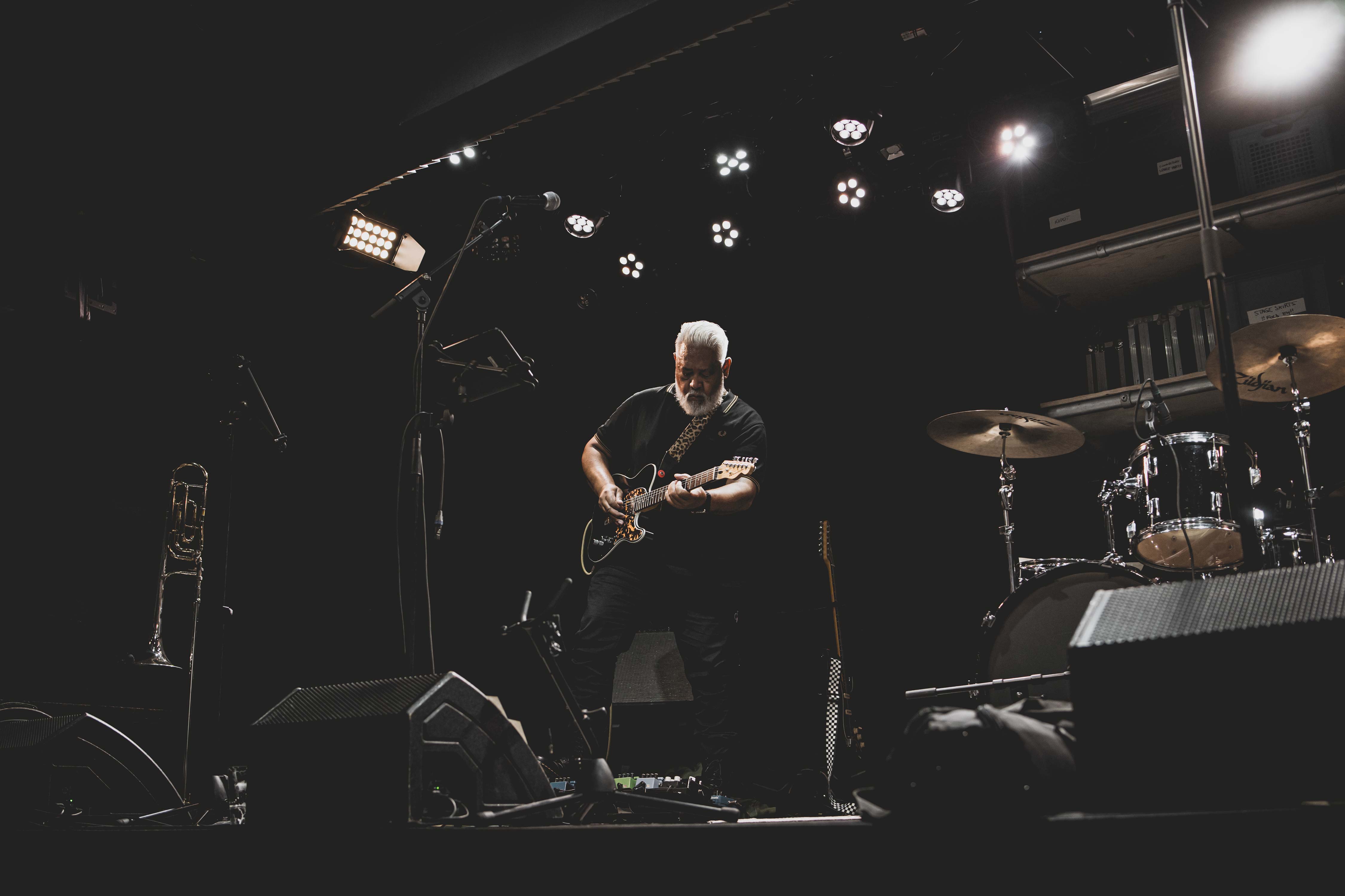 Portret van mannelijke gitarist met grijze baard en custom gitaar op een donker podium, met drumstel en trombone op de achtergrond